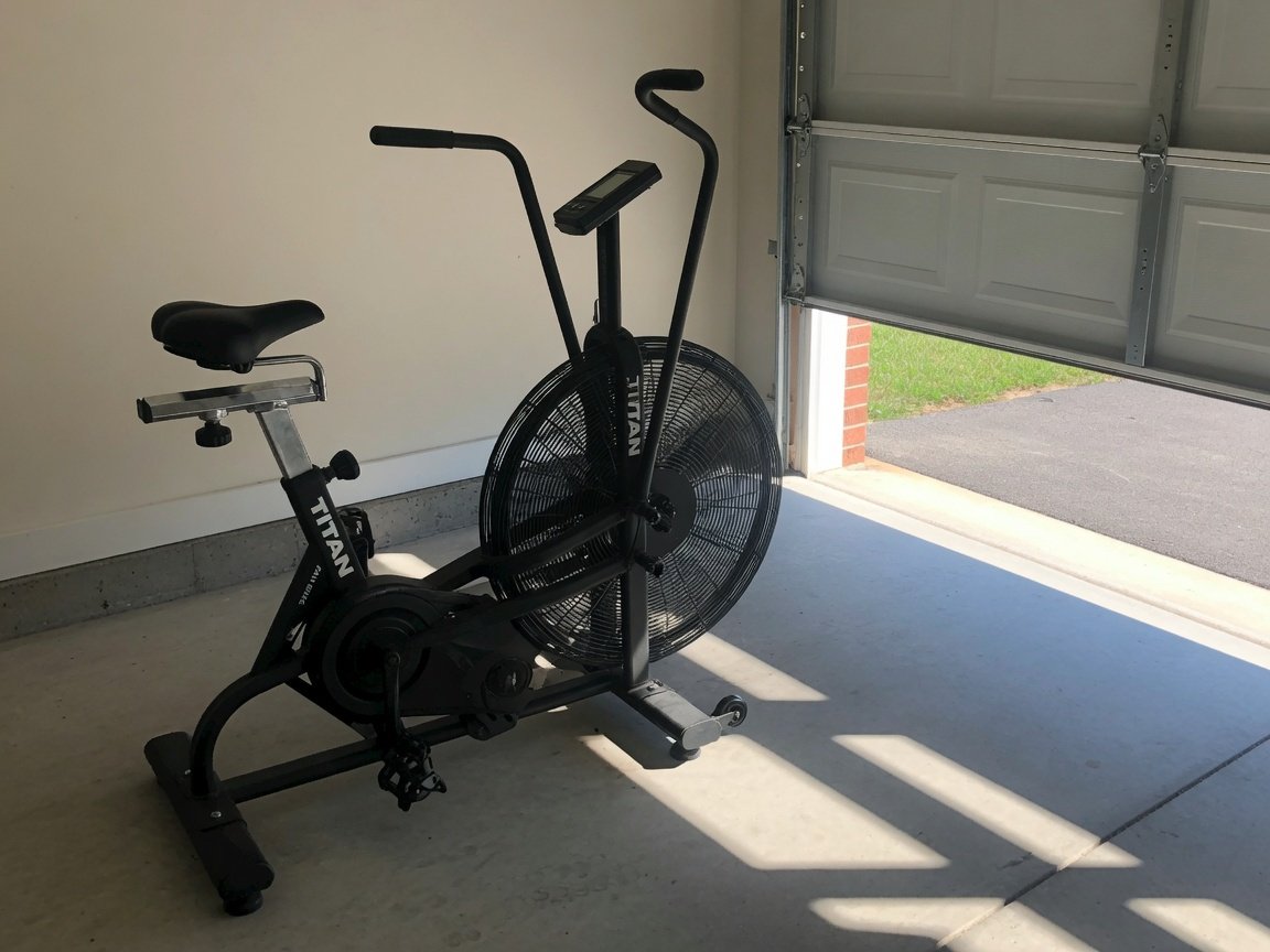 Titan Fitness Fan Bike in matte black positioned beside a half-open garage door, natural daylight spilling in from